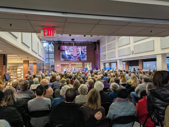 Senator McMorrow speaking onstage as a large crowd in the lobby of the library looks on